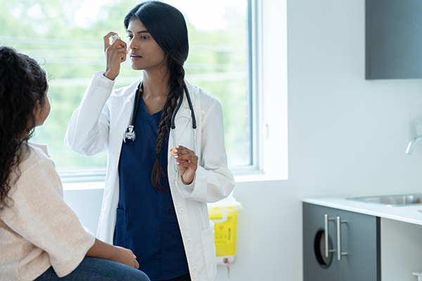 A female doctor holds out an inhaler as she instructs her young patient on how to properly use it for her Asthma. The patient is dressed casually and sitting up on the table as she pays close attention to the instructions.